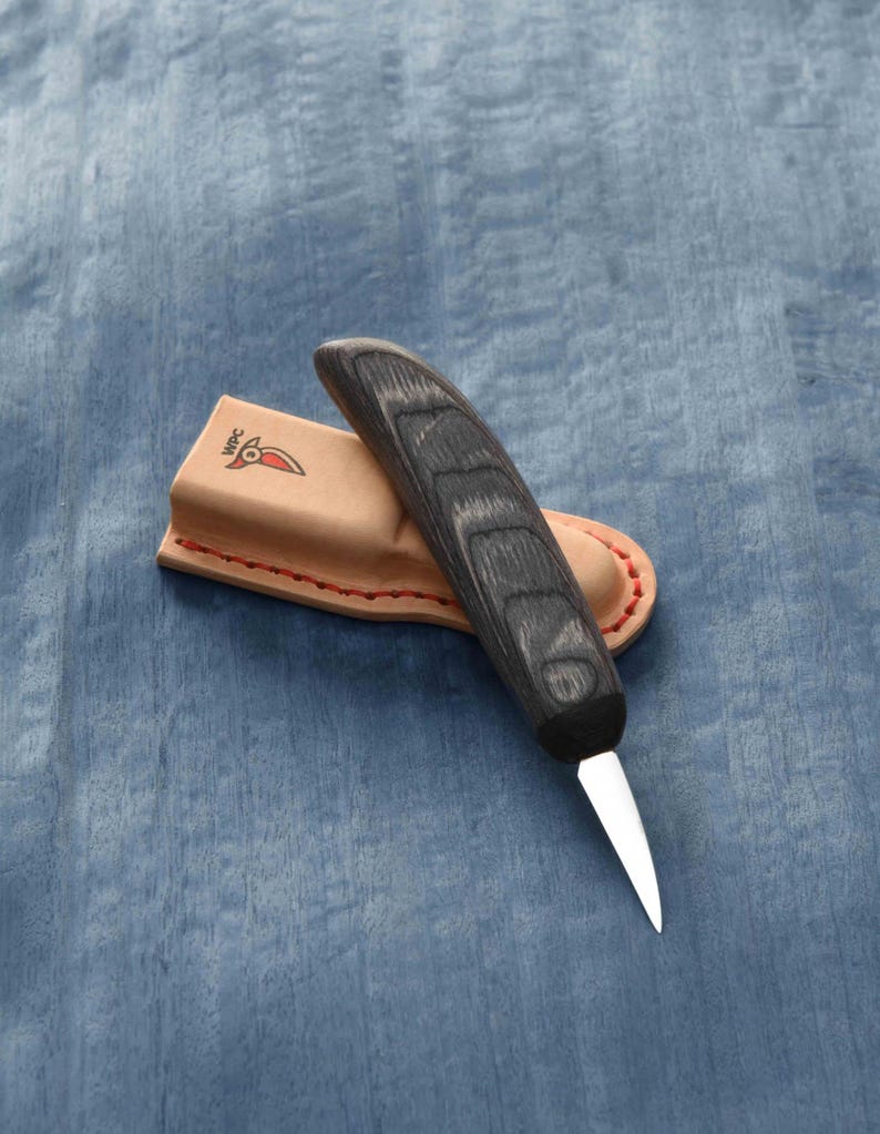 May include: A small, black handled carving knife with a leather sheath. The knife has a curved blade and a wooden handle with a dark wood grain.