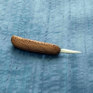 May include: A small, wooden-handled carving knife with a sharp, pointed blade. The handle is made of dark wood with a light brown grain pattern.