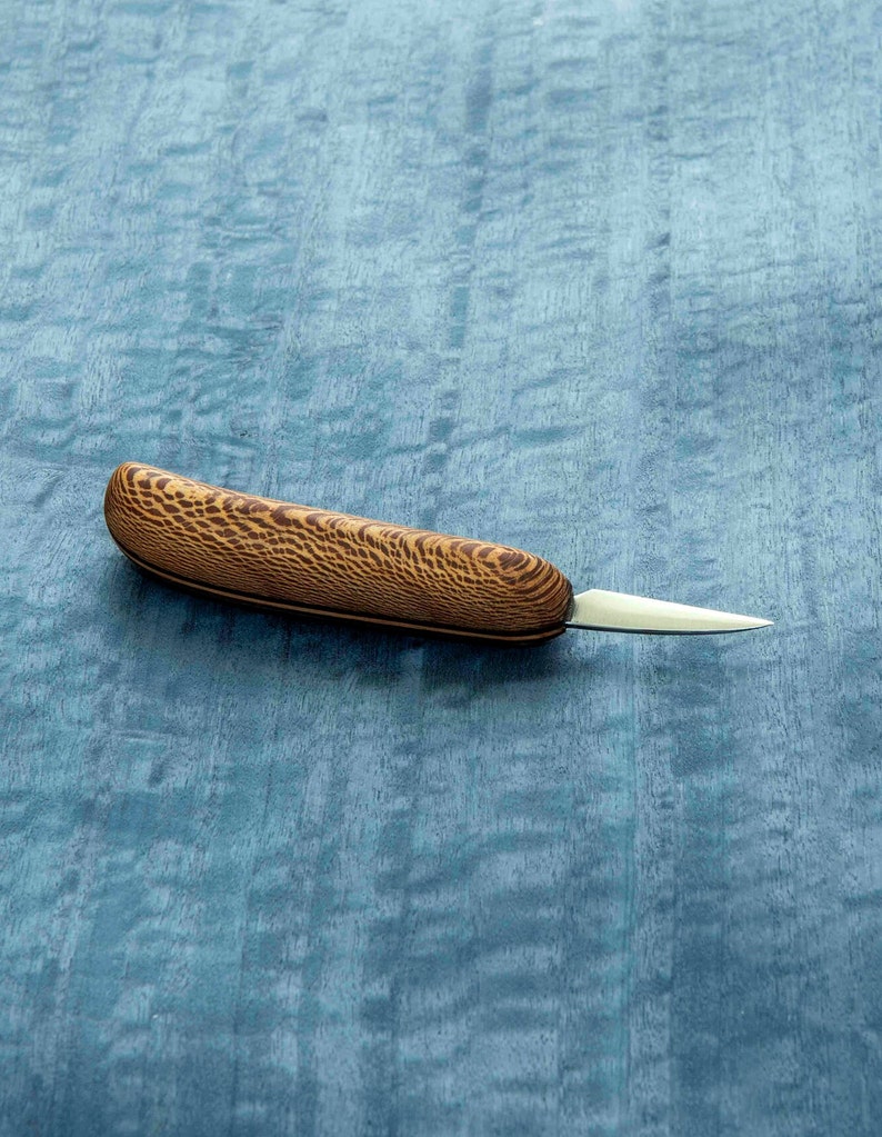 May include: A small, wooden-handled carving knife with a sharp, pointed blade. The handle is made of dark wood with a light brown grain pattern.