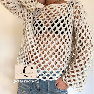 Può includere: Top all'uncinetto color crema a trama aperta con maniche lunghe. Il top ha un design ampio e arioso con scollatura rotonda. Il testo "Clacrochet" e "@clacrochet" sono visibili. La modella indossa jeans blu.