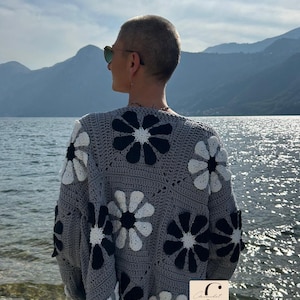 May include: A gray crocheted cardigan with a black and white daisy pattern. The cardigan has a relaxed fit and long sleeves.
