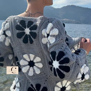 May include: A gray crocheted cardigan with a floral pattern in black and white. The cardigan has a relaxed fit and a slightly cropped length. The cardigan is shown being worn by a person with short hair.