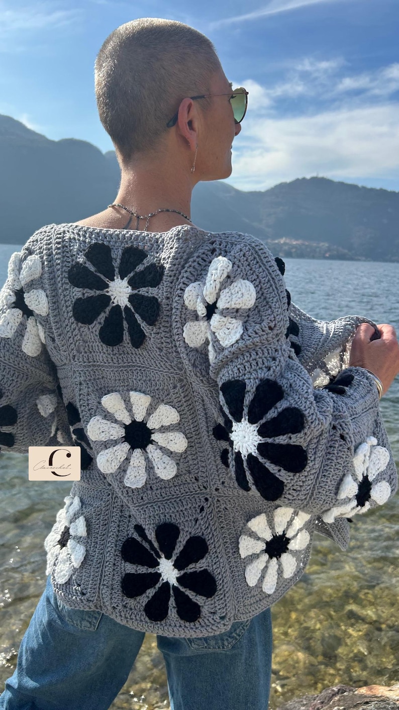 May include: A gray crocheted cardigan with a floral pattern in black and white. The cardigan has a relaxed fit and a slightly cropped length. The cardigan is shown being worn by a person with short hair.
