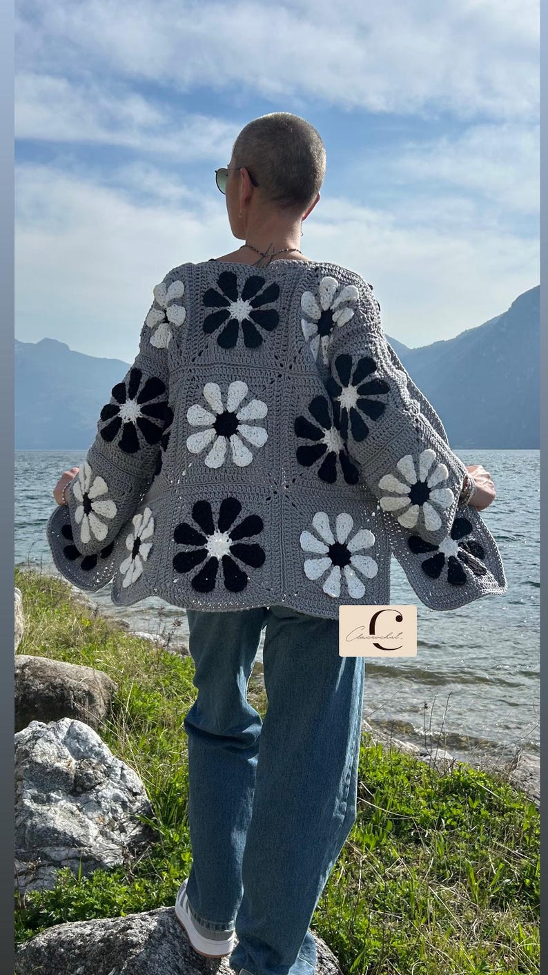May include: A gray crocheted cardigan with a floral pattern in black and white. The cardigan has a relaxed fit and is shown being worn by a person with short hair. The person is standing on a rock by a body of water.