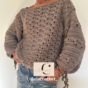 May include: A taupe-colored, handmade crochet sweater with a boat neck and long sleeves. The sweater has a textured, open weave design. The sleeves are gathered at the wrist, and the bottom hem has decorative tassels. The image also includes the text "Clacrochet" and "@clacrochet_."