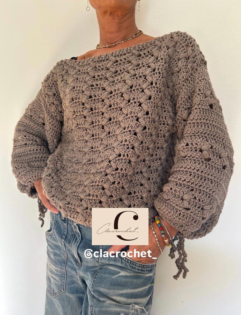 May include: A taupe-colored, handmade crochet sweater with a boat neck and long sleeves. The sweater has a textured, open weave design. The sleeves are gathered at the wrist, and the bottom hem has decorative tassels. The image also includes the text "Clacrochet" and "@clacrochet_."