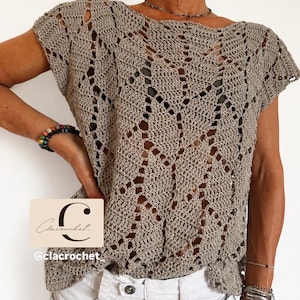 May include: A beige crochet top featuring an openwork leaf pattern, short sleeves, and a wide neckline. The garment is layered over a dark undergarment and paired with white trousers. A logo with the text Clacrochet is visible on the lower left.