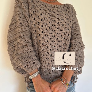 May include: A taupe-colored crochet sweater with a boat neck and long, voluminous sleeves. The sweater features a textured stitch pattern and is worn with bracelets and jeans. The image includes the text "Clacrochet" and the Instagram handle "@clacrochet_".