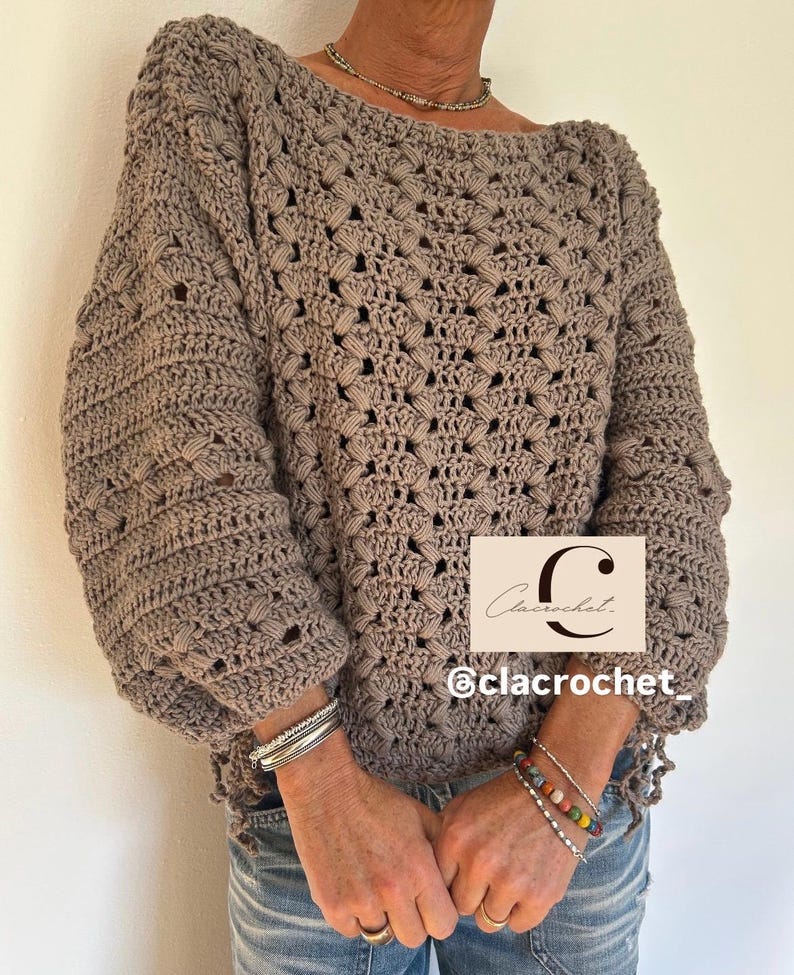 May include: A taupe-colored crochet sweater with a boat neck and long, voluminous sleeves. The sweater features a textured stitch pattern and is worn with bracelets and jeans. The image includes the text "Clacrochet" and the Instagram handle "@clacrochet_".