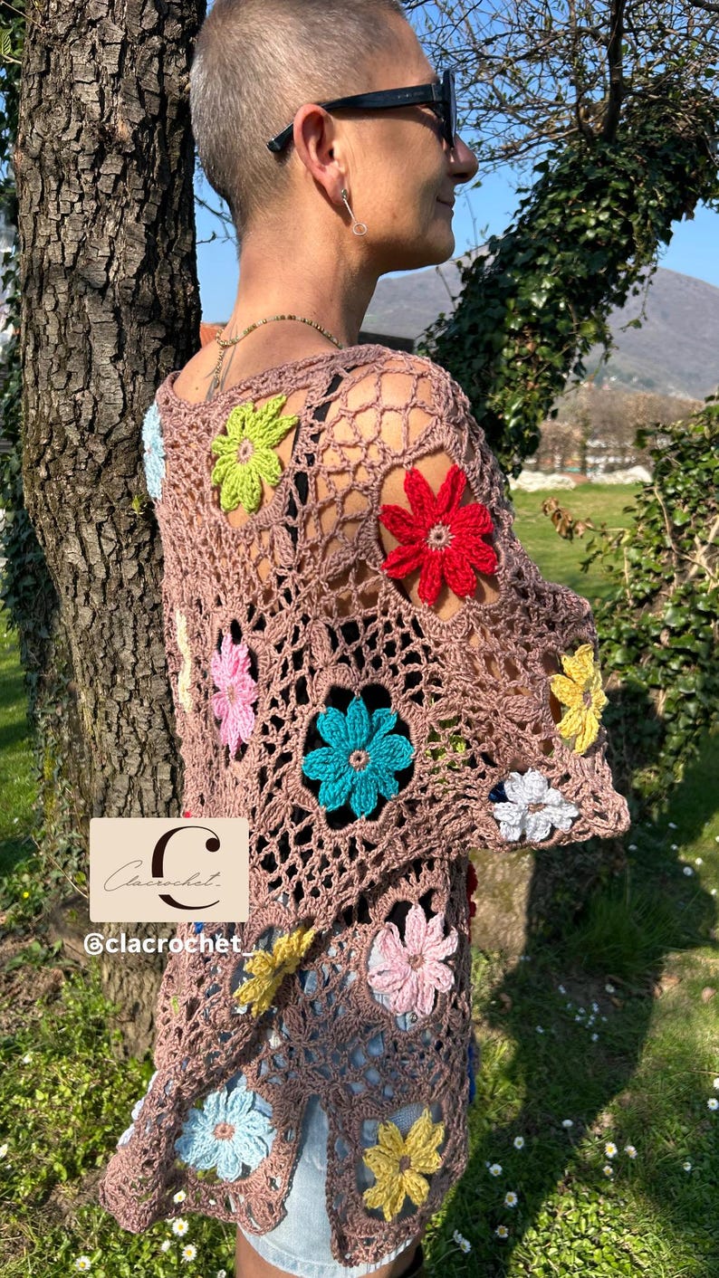 May include: A brown crochet top with colourful, three-dimensional flower appliqu&eacute;s. The open-weave design features flowers in shades of red, yellow, blue, pink, green, and white. The Etsy shop name "clacrochet" is visible in the lower left corner.