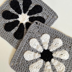 May include: Two crocheted squares with a flower design. One square has a black flower on a gray background. The other square has a white flower on a gray background.