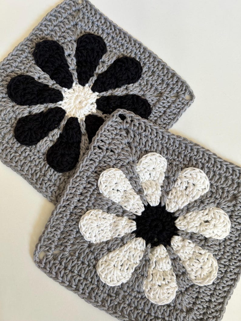 May include: Two crocheted squares with a flower design. One square has a black flower on a gray background. The other square has a white flower on a gray background.