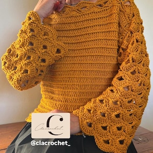 Pode incluir: Top de crochê amarelo mostarda com mangas vazadas e decote com vieiras. O top apresenta um corpete canelado e mangas flare com um padrão geométrico. A marca "Clacrochet" é visível na imagem.