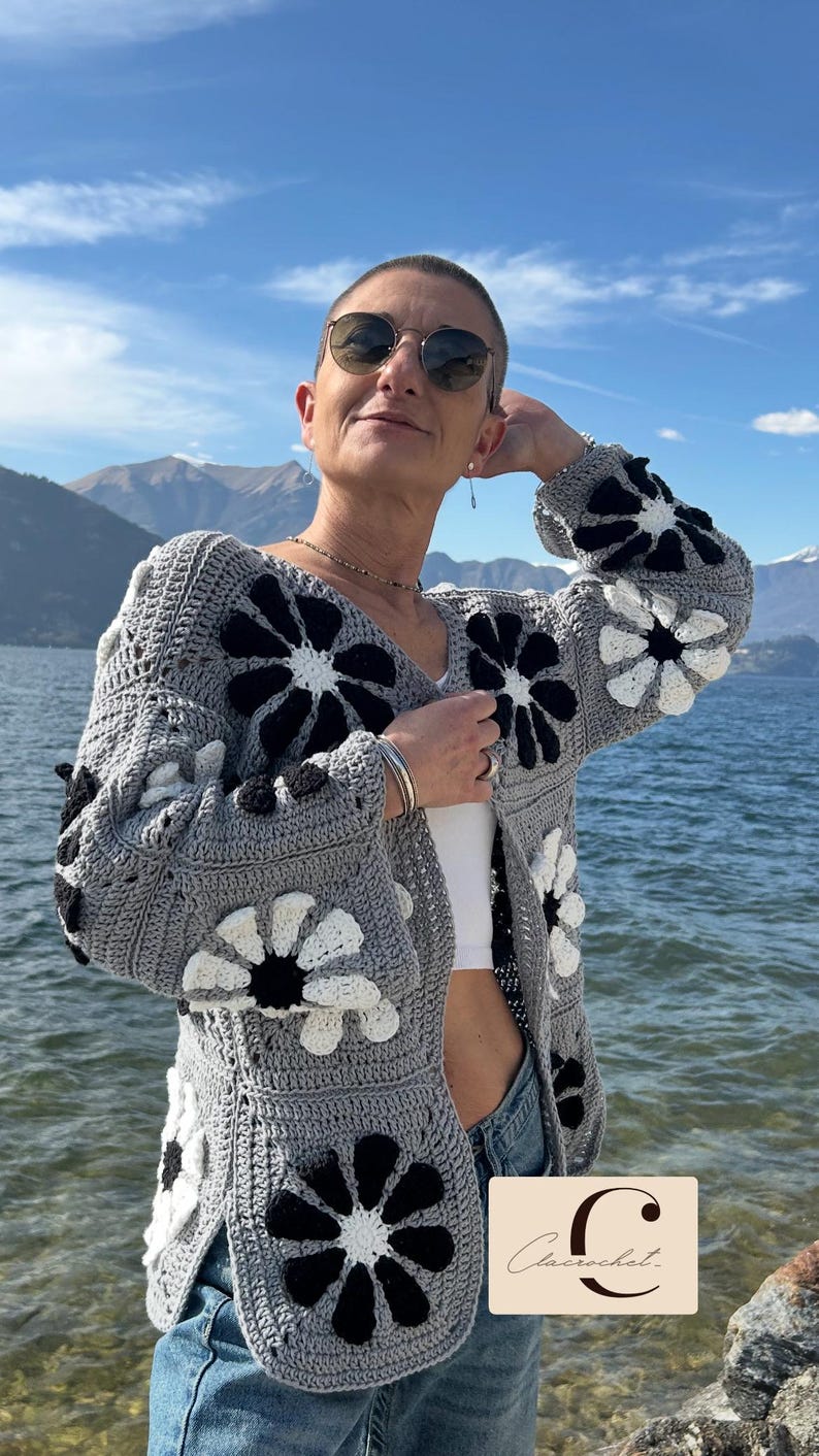 May include: A gray crocheted cardigan with black and white flower designs. The cardigan is open in the front and has a V-neck. The person wearing the cardigan is standing in front of a body of water and mountains in the background. The text "Clacrochet" is visible in the lower right corner of the image.