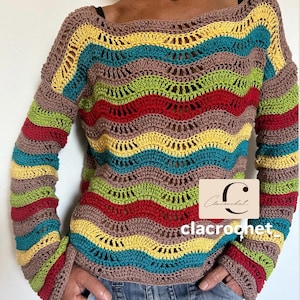 Può includere: Un maglione all'uncinetto a maniche lunghe e multicolore con un motivo a righe nei toni del marrone, giallo, verde, rosso e blu. Il maglione ha uno scollo tondo e maniche svasate. Il testo "clacrochet" è visibile in basso a destra.