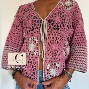 Può includere: Un cardigan all'uncinetto con motivo a piastrelle granny rosa e beige, scollo a V e chiusura frontale con laccetti. Il capo presenta maniche a tre quarti e un bordo beige chiaro. Un logo con la scritta Clacrochet è visibile sul davanti.