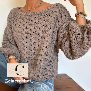 May include: A taupe-colored crochet sweater with a wide neckline and voluminous sleeves. The sweater features a textured stitch pattern. The image also includes the text "Clacrochet" and "@clacrochet_" in a cursive font.