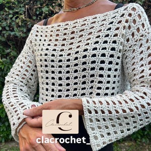 Può includere: Top all'uncinetto bianco sporco con maniche lunghe e design a trama aperta. Il top ha uno scollo a barchetta e maniche a campana. Il nome del marchio "clacrochet" è visibile su una piccola etichetta rettangolare.