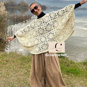 May include: Cream-colored crochet poncho with a floral square pattern. The poncho is worn over a black turtleneck and paired with brown wide-leg pants. The image includes the text "Clacrochet" and "@clacrochet_."