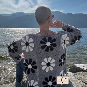 May include: A gray crocheted cardigan with a black and white daisy pattern. The cardigan is worn by a person with short hair, standing on a rocky shore by a body of water.
