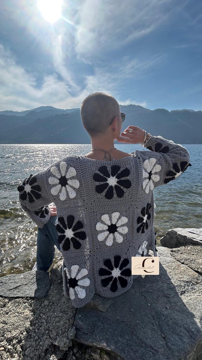 May include: A gray crocheted cardigan with a black and white daisy pattern. The cardigan is worn by a person with short hair, standing on a rocky shore by a body of water.