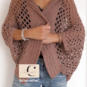 Può includere: Un cardigan all'uncinetto rosa polveroso con cappuccio e design a trama aperta. Il cardigan ha maniche lunghe e viene indossato sopra un top nero e jeans blu. L'immagine include un logo che dice "Clacrochet" e l'handle di Instagram "@clacrochet_".