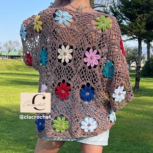 May include: A brown crochet top with colourful flower appliqu&eacute;s. The open-weave design features flowers in red, blue, green, and pink. The top is worn with denim shorts and brown boots, creating a bohemian aesthetic.