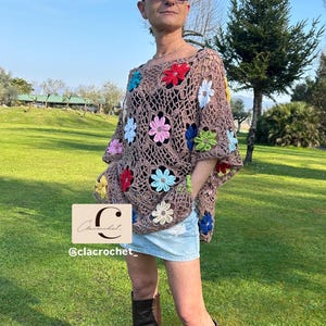 May include: A brown crochet poncho with colourful flower accents. The poncho features a floral pattern with red, blue, pink, and yellow flowers. The image also includes a denim skirt and brown boots. The text "C lacrochet" and "@clacrochet_" is visible.