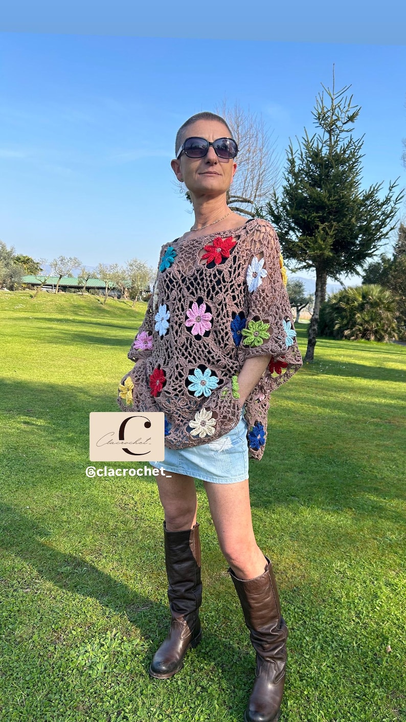 May include: A brown crochet poncho with colourful flower accents. The poncho features a floral pattern with red, blue, pink, and yellow flowers. The image also includes a denim skirt and brown boots. The text "C lacrochet" and "@clacrochet_" is visible.
