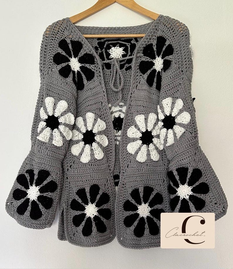 May include: Gray crochet cardigan with black and white flower designs. The cardigan has a tie closure at the neck and bell sleeves.