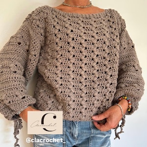May include: A taupe crochet sweater with a boat neck and long sleeves. The sweater has a textured, openwork design and is worn with blue jeans. The sleeves have decorative ties at the cuffs. The image includes a logo that says "Clacrochet" and the Instagram handle "@clacrochet_".