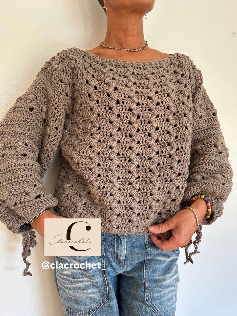 May include: A taupe crochet sweater with a boat neck and long sleeves. The sweater features a textured, openwork pattern and is paired with blue jeans. The sleeves have decorative ties at the cuffs. The image includes a logo that says "Clacrochet" and the Instagram handle "@clacrochet_".