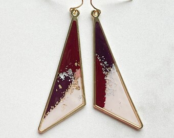 Könnte beinhalten: Ein Paar goldfarbene Ohrringe in Dreiecksform. Die Ohrringe sind mit einem weißen, burgunderroten und violetten Harz gefüllt, das mit goldenen Glitzerakzenten verziert ist.