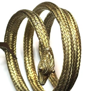 Pode incluir: Uma pulseira de metal dourado em forma de serpente enrolada. O corpo apresenta uma textura de malha trançada flexível, enquanto a cabeça é detalhada com escamas gravadas e pequenos olhos escuros incrustados em uma formação espiral.