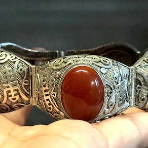 Pode incluir: Uma pulseira articulada com três grandes pedras ovais de cor vermelho-escura, lisas, engastadas numa intrincada estrutura de metal prateado. Os segmentos metálicos apresentam padrões de filigrana detalhados e caracteres chineses em relevo.
