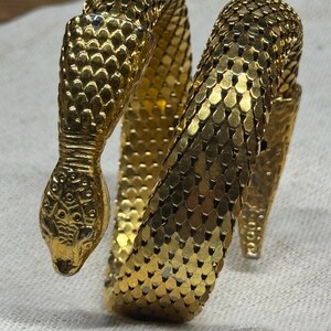 Könnte beinhalten: Ein goldfarbenes Metallarmband in Form einer gewundenen Schlange. Der Körper des Armbands weist ein flexibles, ineinandergreifendes Schuppenmuster auf, während der Kopf mit eingravierten Markierungen und kleinen, dunklen Augen versehen ist.