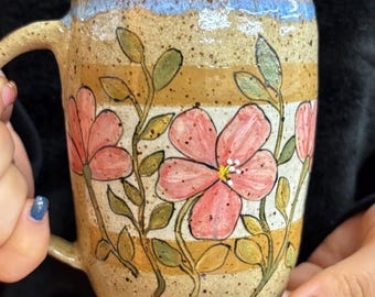 May include: A ceramic mug with a speckled beige finish, featuring a hand-painted design of pink flowers with green stems and leaves. The mug has horizontal tan stripes and a blue-tinted rim, with a curved handle extending from the side.