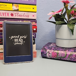 May include: A black and blue Kindle e-reader with a white screen displaying the text "good girls + read + smut." The e-reader is sitting on a table with a stack of books behind it and a potted plant with pink flowers to the right.