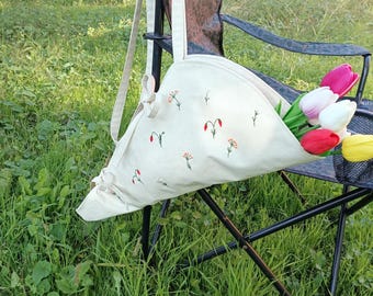 May include: A cream-colored canvas tote bag with a triangular shape and long shoulder straps, featuring small embroidered floral motifs in red, orange, and green. The bag contains a bouquet of artificial tulips in red, white, and yellow.