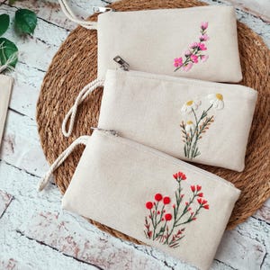 Op de afbeelding: Drie beige canvas pouches met geborduurde bloemmotieven. Eén pouch heeft roze bloemen, een andere witte madeliefjes en de derde rode bessen en bloemen. Elke pouch heeft een ritssluiting en een witte polsband.