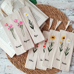 May include: A collection of embroidered fabric bookmarks featuring floral designs. Each bookmark has a different flower and name embroidered on it. The bookmarks are beige with white tassels and are arranged on a woven mat with an open book.