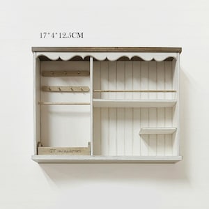 Pu&ograve; includere: Una mensola da parete in legno bianco con una parte superiore smerlata e un bordo marrone scuro. &Egrave; dotata di pi&ugrave; scomparti, tra cui ganci, un ripiano e una piccola scatola. La mensola misura 17cm x 4cm x 12,5cm.