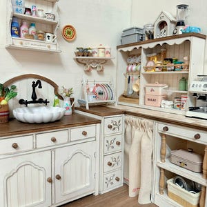 Pu&ograve; includere: Una scena di cucina in miniatura con mobili bianchi, un lavandino a vasca e un piano di lavoro in legno. La scena comprende mensole con utensili da cucina in miniatura, un fornello e una piccola scatola per il pane. Il testo "Display only. Props not included" &egrave; visibile.