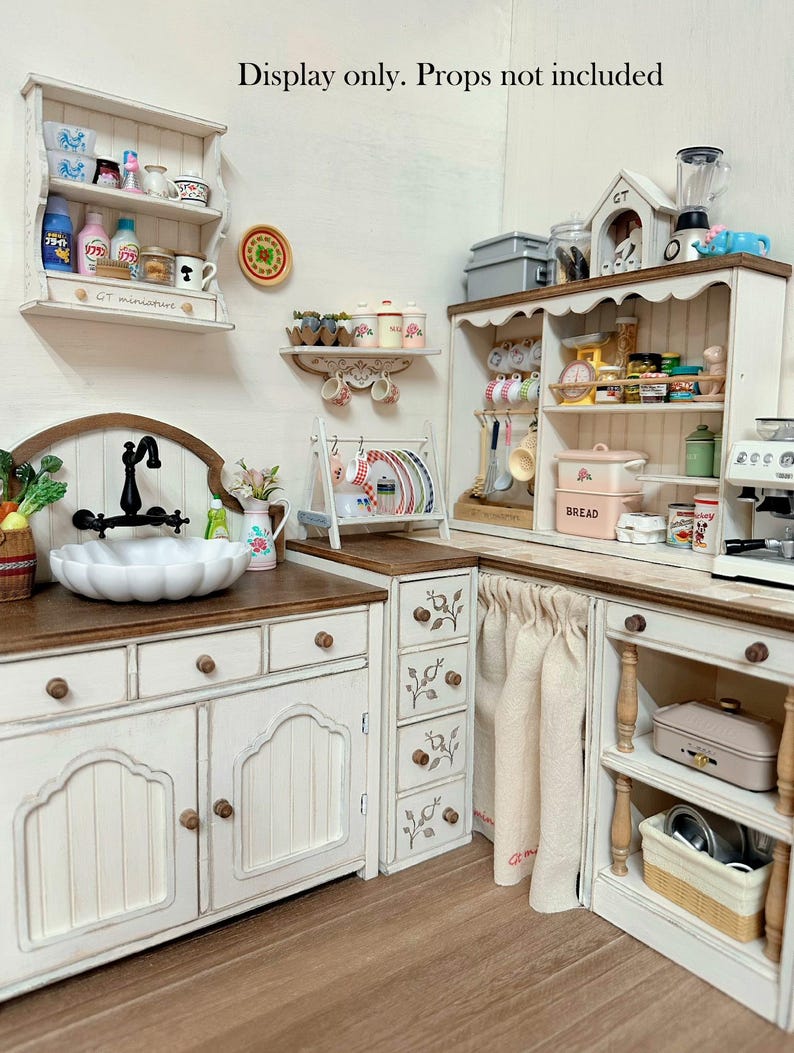 Pu&ograve; includere: Una scena di cucina in miniatura con mobili bianchi, un lavandino a vasca e un piano di lavoro in legno. La scena comprende mensole con utensili da cucina in miniatura, un fornello e una piccola scatola per il pane. Il testo "Display only. Props not included" &egrave; visibile.