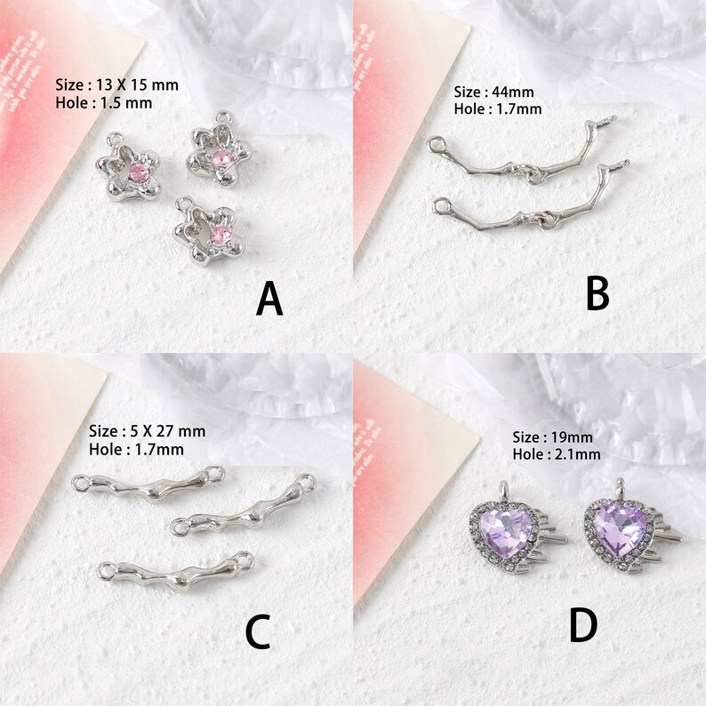 May include: Four different jewelry findings with various sizes and shapes.  The first finding is a silver flower with a pink rhinestone center. The second finding is a silver connector with two loops. The third finding is a silver connector with three loops. The fourth finding is a silver heart with a purple rhinestone center.