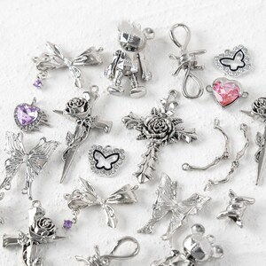 May include: A collection of silver-toned charms featuring various designs, including butterflies, hearts, roses, crosses, and a robot. The charms are scattered on a white surface.
