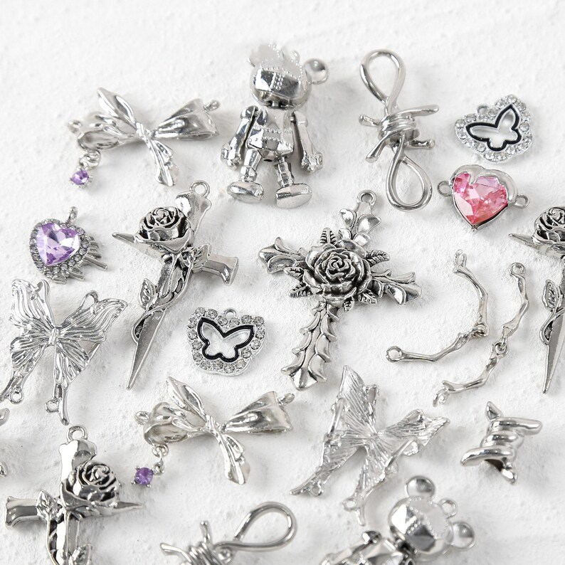 May include: A collection of silver-toned charms featuring various designs, including butterflies, hearts, roses, crosses, and a robot. The charms are scattered on a white surface.