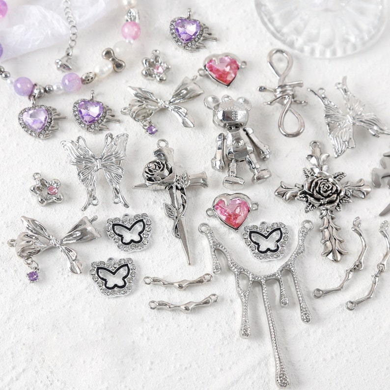 May include: Assortment of silver-toned metal charms with various designs, including butterflies, bows, hearts, crosses, and a robot. Some charms feature pink and purple rhinestones.