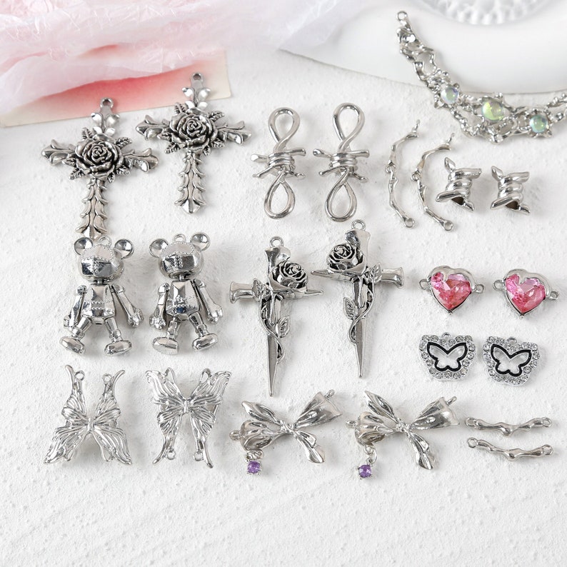May include: A collection of silver-toned metal charms featuring various designs, including crosses with roses, barbed wire, butterflies, bows, hearts, and teddy bears.