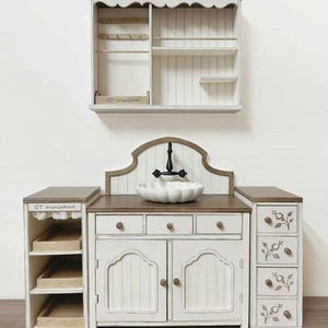 Pu&ograve; includere: Un set da cucina in miniatura bianco e marrone con lavandino, armadietti e una mensola a muro. Il lavandino ha una ciotola bianca smerlata e un rubinetto nero. Gli armadietti hanno cassetti e ante con decorazioni floreali. La mensola ha ganci e piccole mensole.